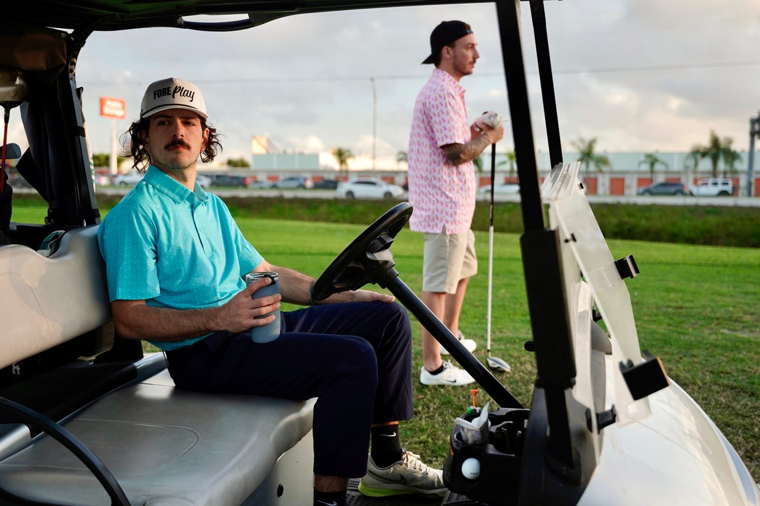 Two golfers at a golf cart, one seated holding a tumbler