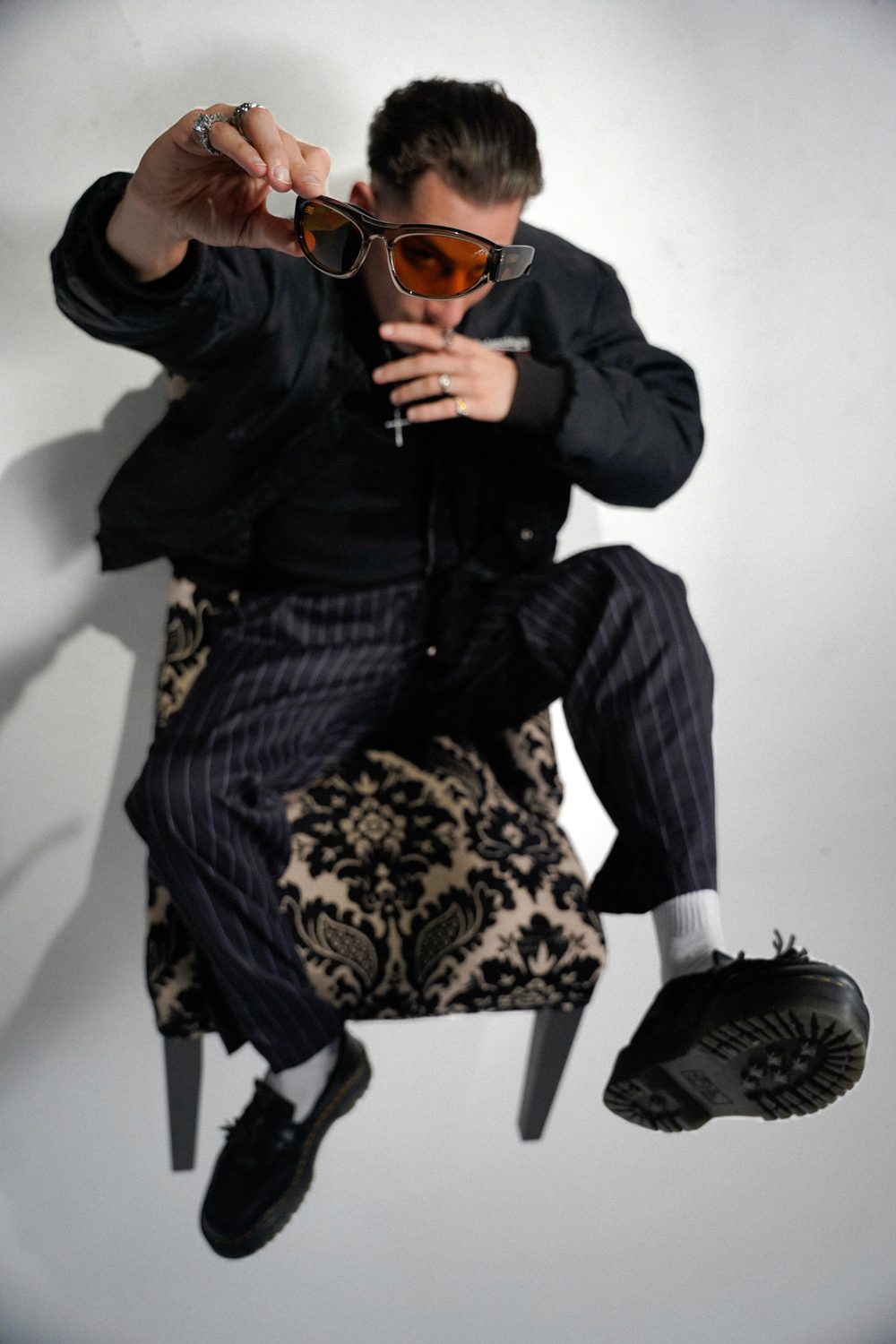Man in a bomber jacket seated on a patterned chair, holding orange-lens sunglasses toward the camera