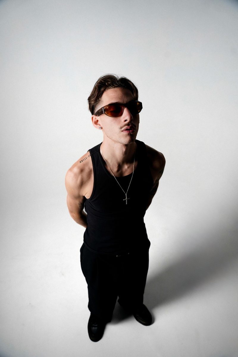 Man in a black tank top and orange-lens sunglasses, photographed from above against a white wall