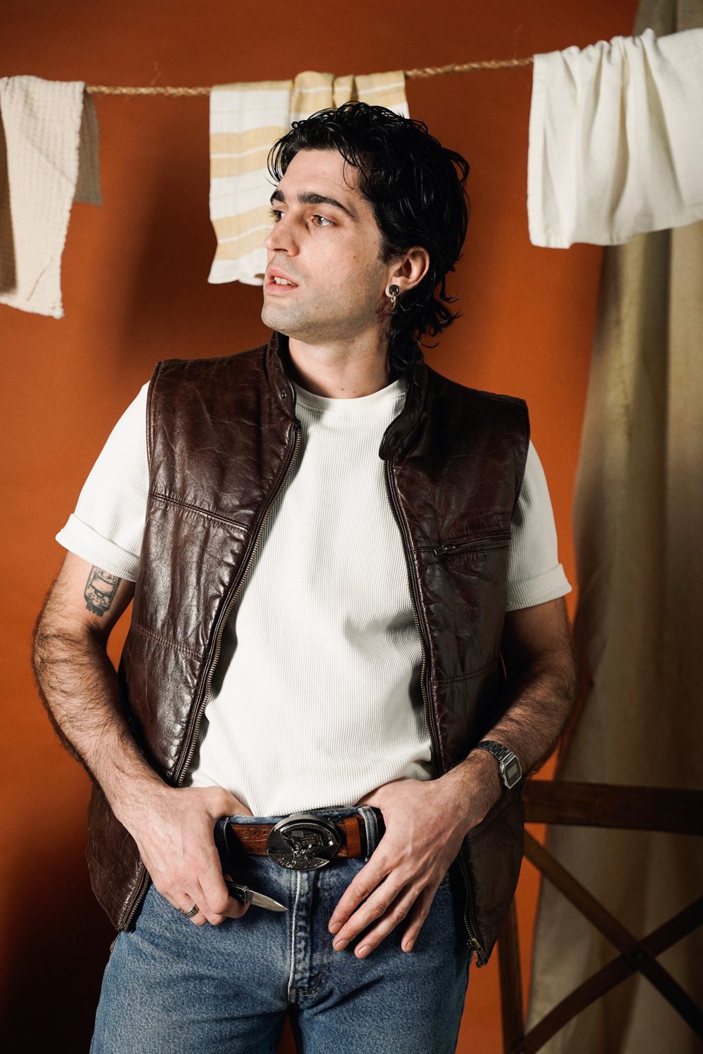 Man in a leather vest and jeans with a western belt buckle against an orange wall