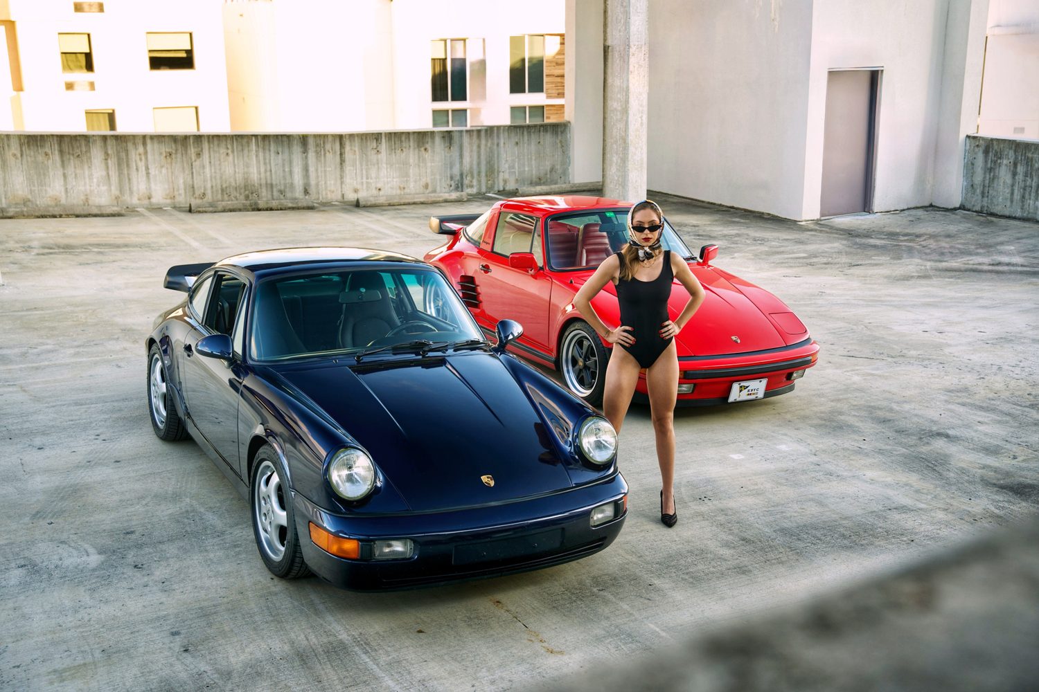 Two classic Porsche 911s, one navy and one red, on a rooftop parking deck with a model standing between them